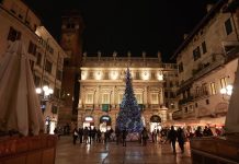 Gli alberi di Natale Bauli, icone natalizie della città di Verona Albero di Natale Bauli acceso in Piazza delle Erbe di notte, circondato dai passanti sotto Palazzo Maffei