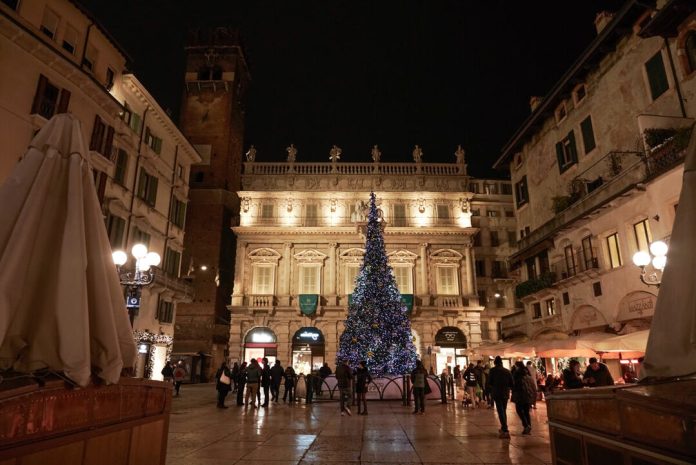Albero di Natale Bauli acceso in Piazza delle Erbe di notte, circondato dai passanti sotto Palazzo Maffei